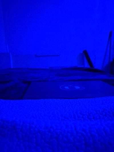This image depicts an indoor bedroom scene in Shoreline, United States of America, entirely illuminated by a deep blue light. In the foreground, a textured, light-colored fuzzy blanket covers the lower third of the frame. Positioned in the midground, a closed laptop computer with a prominent, circular two-part logo on its lid rests on a dark-colored surface, likely a bed. The background consists of a plain wall, also bathed in blue light. A narrow, horizontal shelf or ledge is affixed to the wall on the left. To the right, indistinct vertical objects, including a dark pole leaning against the wall and a cylindrical container, are partially visible. No individuals are present, and no actions are occurring.