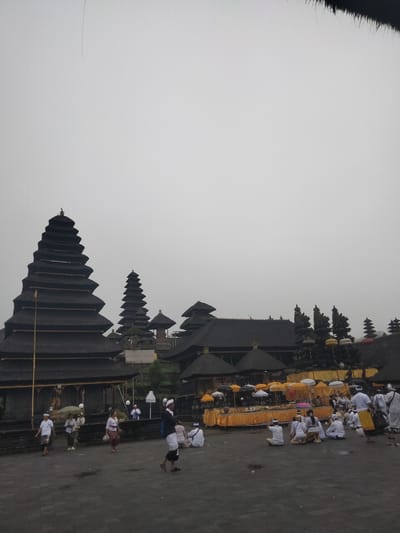 The image depicts a temple complex in Rendang, Indonesia on a cloudy day. The structures are built in traditional Balinese style with tiered roofs and dark materials. A large group of people, mostly dressed in white ceremonial attire, are gathered in a courtyard. Some are walking, others are sitting on the ground. Offerings, including colorful umbrellas and decorations, are visible on a raised platform. The ground is paved with stone. The overall atmosphere suggests a religious ceremony or gathering.