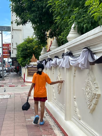 A young person with dark hair, wearing an orange t-shirt and dark shorts, walks away from the camera on a red-tiled sidewalk. They are carrying a black plastic bag in their left hand and their right arm is extended towards a decorative white wall. The wall has ornate carvings and is draped with fabric in black and white. The background shows a street scene with a building, trees, and a cloudy sky. A sign in Thai is visible on the building. A pigeon is on the sidewalk in the foreground. The location is identified as Surat Thani City Municipality, Thailand.