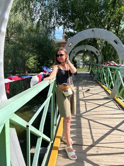 A woman with long brown hair, wearing sunglasses, a black tank top, khaki shorts, and white sandals, stands on a wooden bridge in Medvedevo Urban Settlement, Russia. She has a small crossbody bag with a circular design. The bridge has a light green railing decorated with red, white, and blue flags. Archways made of gray metal extend over the bridge. Lush green trees and foliage surround the area. Sunlight casts shadows on the wooden planks of the bridge. A bird is visible on the bridge in the background. The water below the bridge is dark and appears to have at least one duck swimming in it.