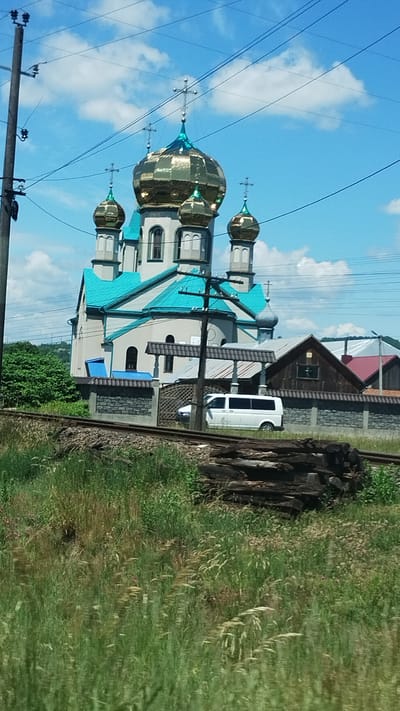This media file depicts a picturesque scene in Teresva, Ukraine, featuring an Eastern Orthodox church with striking golden domes and a vibrant turquoise roof. The church is situated next to a railway track, with piles of wood and lush green grass flanking the tracks, suggesting a rural or semi-rural setting. A white van is parked near the church, and the sky above is a brilliant blue with scattered white clouds, indicating pleasant weather, likely during the daytime. Overhead power lines and a utility pole are visible in the foreground, adding to the sense of a lived-in landscape. The overall impression is one of tranquility and beauty, with the sunlight reflecting off the church's golden domes. There is no visible text in the image.