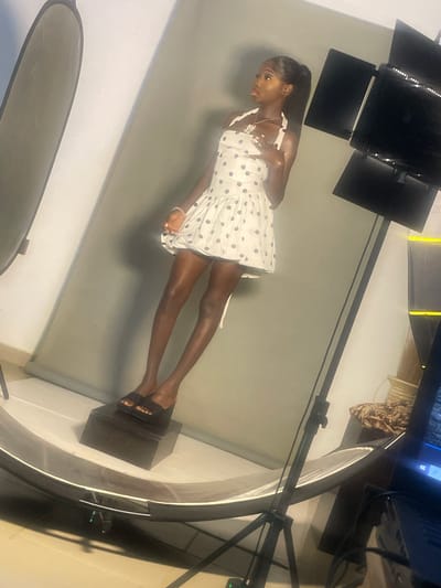 A young Black woman is posing for a photo shoot in a studio. She is wearing a white dress with black polka dots and black sandals. Her hair is pulled back in a ponytail, and she is wearing a necklace and bracelet. She is standing on a dark rectangular platform.
The studio has a light gray backdrop. There are studio lights set up around the woman, including a large ring light. One of the lights is positioned above and to the left of the woman, with multiple panels. Another light is visible to the right of the scene, appearing to be part of a larger lighting setup. In the background, there is a glimpse of a monitor screen and some decorative items, including a patterned fabric.
The lighting suggests it is daytime, but the indoor setting makes it independent of the actual time of day or weather outside. The woman appears focused and poised, likely engaged in modeling or a similar professional photo session. The overall scene is professional and well-lit, typical of a studio photography environment. The image is oriented sideways. There is no visible text.