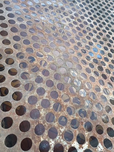 This media file captures a close-up, high-angle view of a rusted metal grate with circular perforations. The grate is tilted, creating a sense of depth and perspective. Through the holes, a patterned surface is visible, suggesting it might be a table or bench top. The pattern appears to be abstract with swirling lines and variations in color, predominantly in shades of brown, beige, and dark gray. The metal itself is weathered, exhibiting patches of rust and a textured surface. The lighting suggests it is daytime, with natural light reflecting off the metal and illuminating the scene. There are no people, animals, or specific events depicted; the focus is purely on the texture and pattern of the object. The location context of Tobyl, Kazakhstan, offers no immediate visual cues within the image itself, as the scene is generic and focuses on material detail.