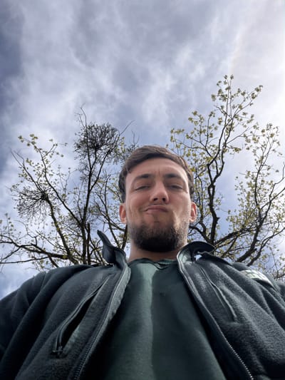A low-angle selfie captures a bearded man in a dark green fleece jacket, positioned against a backdrop of a cloudy sky and tree branches with budding leaves. The man's face is the focal point, with the camera angled upwards. The location appears to be outdoors. There is no indication of the location of Kosti, Bulgaria, being in the image.