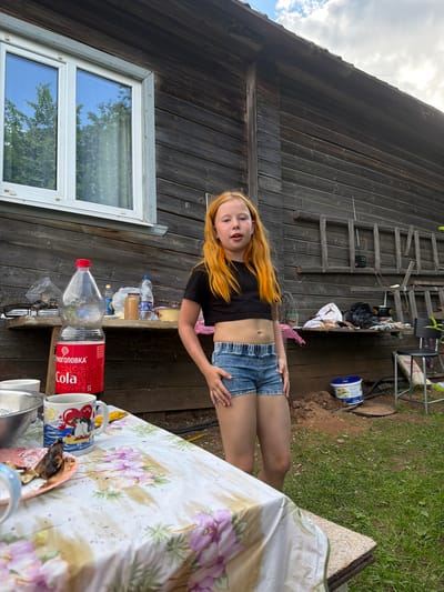 A young girl with long, bright orange hair and fair skin stands in front of a rustic wooden building. She is wearing a black crop top and denim shorts. The building appears to be a shed or small house, with weathered wooden planks and a window with white trim. A ladder leans against the side of the building, and various items, including buckets and bags, are scattered around. In the foreground, a table is set with a floral tablecloth, mugs, and a large bottle of "Cola" with Russian text. The scene suggests an outdoor setting, possibly a backyard or rural area, with green grass and dirt visible. The lighting indicates it is daytime, with the sun casting shadows. The girl's expression is neutral, and she is looking towards the camera. The overall impression is a casual, outdoor scene. The visible text on the bottle reads " Cola" and "СУПЕРКОЛО".