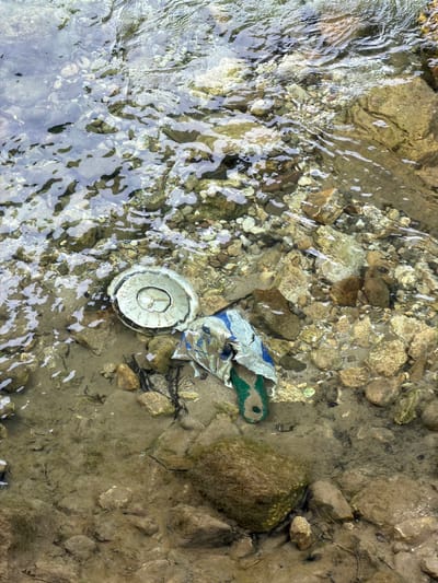 The image depicts a section of a shallow, rocky stream bed in Konjic, Bosnia and Herzegovina. The water is clear, allowing visibility of the submerged stones and a small amount of debris. The primary objects of interest are two pieces of discarded refuse: a crumpled, torn plastic wrapper, predominantly white with blue and green elements, and a circular, discolored lid with a concentric ring pattern and a small symbol resembling a peace sign in the center. The lid appears to be made of plastic or a similar material and is slightly eroded.
The scene is likely a natural environment, possibly a river or creek, given the presence of flowing water and the rocky substrate. The lighting suggests it is daytime, with natural light reflecting off the water's surface, creating shimmering patterns and obscuring some details of the stream bed. No people are visible in the image. The overall impression is one of environmental neglect, with man-made waste polluting a natural watercourse. There is no visible text in the image.