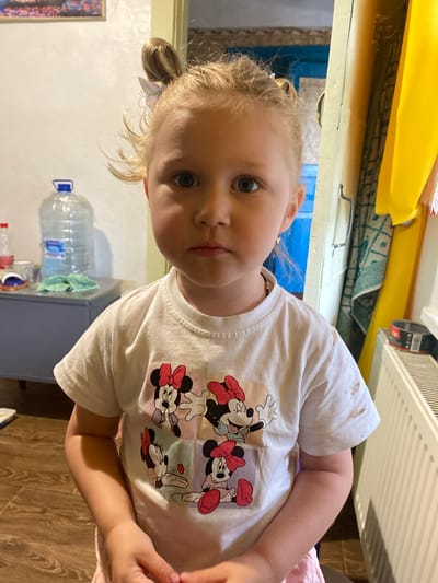 The media file shows a close-up, slightly angled portrait of a young blonde girl indoors. She is wearing a white t-shirt featuring a graphic of Minnie Mouse characters. The girl has light-colored hair pulled back loosely, with a small white bow. Her expression is neutral, and she is looking towards the camera.

Behind the girl, the background includes household items. To the left, a yellow object is visible, possibly a piece of furniture or decoration. Below that, a blue doorframe or wall section can be seen. To the right of the girl, a white ribbed radiator or vent is prominent. In the lower portion of the image, a large clear plastic bottle of water with a blue cap is present, along with other unidentifiable objects on what appears to be a table or shelf. The floor is a wooden laminate.

The lighting suggests an indoor setting, likely during the daytime, as there are no strong indications of artificial lighting. The overall scene appears to be a casual, domestic environment. No specific activity is depicted other than the girl posing for the photograph. The image is sharp and in focus on the girl's face and torso.