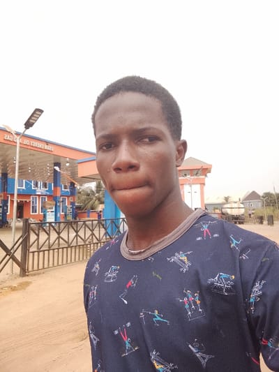 A young male subject with dark skin and closely-cropped dark hair is positioned in the foreground, looking directly at the camera with a neutral expression and slightly pursed lips. He wears a dark blue t-shirt featuring a grey collar and a repeating pattern of multicolored stylized figures in various athletic poses, along with architectural designs.
The subject stands on an unpaved, sandy ground. Behind him, a light brown wooden fence with a criss-cross lattice design separates the foreground from the background. To the left, a metal pole with a modern solar-powered street light is visible.
The background features a gas station structure, predominantly blue with orange accents and white window frames, under a canopy displaying the inverted text "USAM ENERGY OIL AND GAS" on a horizontal orange sign. Multiple fuel pumps are discernible beneath the canopy, and several indistinct individuals are present within the station grounds. Further to the right, a white tanker truck is parked, and distant residential buildings with dark roofs are visible among palm trees. The sky is bright and uniformly overcast. The scene is located in Awkuzu, Nigeria.