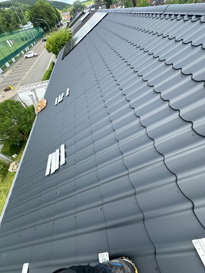 The image provides an elevated view of a dark gray, tile-profiled metal roof, likely undergoing installation or maintenance, in Theux, Belgium. The roof panels are secured with numerous screws. A person's right foot, wearing a dark blue and yellow boot with "V-RISK" text visible on the side, is positioned on the roof in the lower right corner, suggesting someone is on the roof.
Several white, metallic mounting brackets are scattered across the roof surface, particularly in the lower half, indicating preparations for attaching additional structures, possibly solar panels, as a section of solar panels is visible on an adjacent roof or roof section further down. A silver ladder leans against the edge of the roof in the lower left, and a cardboard box sits nearby.
The surrounding environment, viewed from the roof, includes a street with a white car and a dark car parked on the side. Beyond the street, there are bright green tennis courts with white lines and nets, flanked by numerous green trees. In the background, other buildings and more extensive green areas are visible under what appears to be bright, possibly overcast, daytime lighting. The perspective is tilted, looking down from the roof.