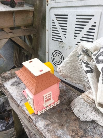 The image displays an outdoor scene in Kon Tum, Vietnam. A pink plastic toy house with a brown tiled roof and a white removable flat roof component rests on a weathered, dark wooden surface. The toy house has white window frames, a white miniature picket fence at the front, and a yellow ramp or step at its base. Behind and to the right of the toy house, a large, light-colored appliance, likely a refrigerator or washing machine, stands vertically, showing a rectangular ventilation grille and a circular fan opening. Adjacent to the appliance, a crumpled white woven sack with visible black lettering is partially obscured. In the upper left background, a rough wooden structure supports a dark object with red elements. The ground beneath the wooden structure contains dark, crumpled material. No individuals or active interactions are present.