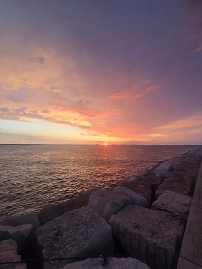 The image depicts a sunset over the Adriatic Sea, viewed from a breakwater composed of large, irregular grey stones. The sky is a gradient of vibrant colors, transitioning from a soft orange and pink near the horizon to a deep purple at the top. Clouds are scattered across the sky, catching the sunset's light. The sea is calm, with gentle ripples reflecting the warm hues of the sky. In the distance, several ships and what appears to be a lighthouse are visible on the horizon. The breakwater extends to the right of the frame, with a paved walkway visible on its far edge. Two fishing rods are positioned on the rocks in the foreground. The scene suggests a peaceful evening by the water, likely in the city of Chioggia, Italy, known for its coastal setting and maritime activities.