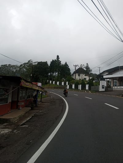The image depicts a curved asphalt road in a semi-rural or mountainous setting, observed on an overcast day. A white solid line marks the left edge of the road, which sweeps to the right. White dashed lines are visible further along the curve, indicating lane separation.
On the immediate left, a single-story structure with red-painted walls and a dark roof serves as a shop, featuring a sign with text that includes "SOUVENIR OLEH-OLEH." A person is partially visible near the entrance of this shop. The shop sits on a dirt and concrete shoulder adjacent to the road, with dense green trees and foliage rising behind it.
On the road, a dark-colored motorcycle with a single rider wearing a dark helmet and clothing is traveling away from the viewer, moving around the curve.
To the right of the road, a landscaped area is bordered by a series of low, white and dark retaining walls or fences. Behind these, a large, multi-story white house with a distinctive dark, possibly tiled or thatched, roof is situated on a higher elevation amidst trees. Further along the road on the right, a modern-style building with a dark roof and white walls, possibly an entrance gate or part of a complex, features a sign reading "MASUK." Overhead, multiple utility lines are visible against the gray sky, supported by power poles.
As per the provided information, the location is the city of Saing Ta Mawt Lay, Myanmar. However, the presence of Indonesian/Malay language on signage ("OLEH-OLEH," "MASUK") suggests a different geographical context, such as Indonesia or Malaysia.