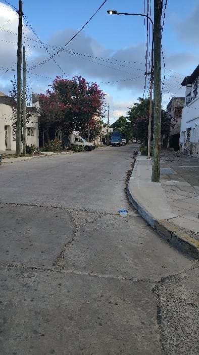 This image depicts a residential street scene in the city of Caseros, Argentina, viewed from a low angle looking down the street. The sky is partially cloudy with patches of blue.
The street is paved with concrete or asphalt, exhibiting multiple cracks and repairs, and appears generally worn. It extends into the distance, with a slight bend to the right. Concrete sidewalks with grey curbs line both sides of the street; the curb on the right side in the foreground has sections painted yellow. A small, light blue plastic object is visible on the sidewalk's edge near the right foreground curb.
Multiple wooden utility poles stand along both sides of the street, supporting a dense network of overhead wires that crisscross the sky. A tall wooden utility pole is prominent on the right side of the street, supporting an illuminated modern LED streetlamp extending over the roadway. Another utility pole is visible on the left side in the foreground.
Residential buildings line both sides of the street. These structures are mostly light-colored, with some showing exposed textured facades. Vegetation, including trees and bushes, is present along the street, partially obscuring some buildings. A prominent tree with dense reddish-pink foliage stands on the left side of the street, mid-ground.
Several vehicles are visible along the street: a white van is parked on the left side in the mid-ground, near the reddish-pink tree. Further down the street, a dark blue truck is parked on the right side. Additional vehicles are discernible in the far distance. No individuals are present in the visible area. The scene is static.