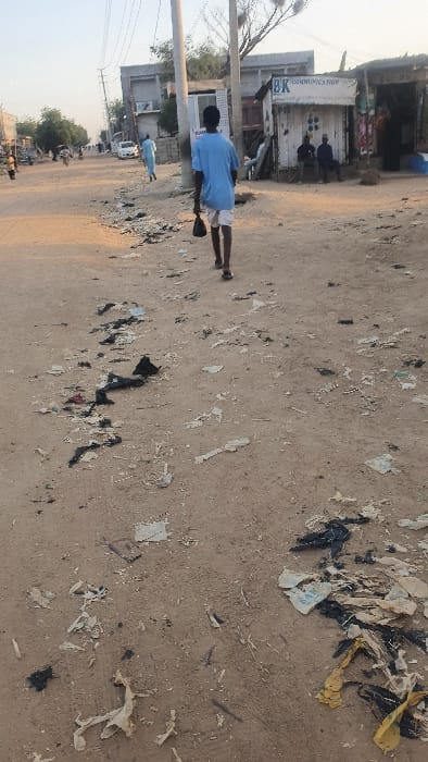 A street scene reveals a dirt road littered with various trash, including plastic bags and paper scraps. A man in a blue shirt and light-colored shorts walks along the road, sweeping the debris. Buildings line one side of the street, with a store visible in the background. Power poles and wires run along the street, and the sky is bright. Other people are seen in the distance.