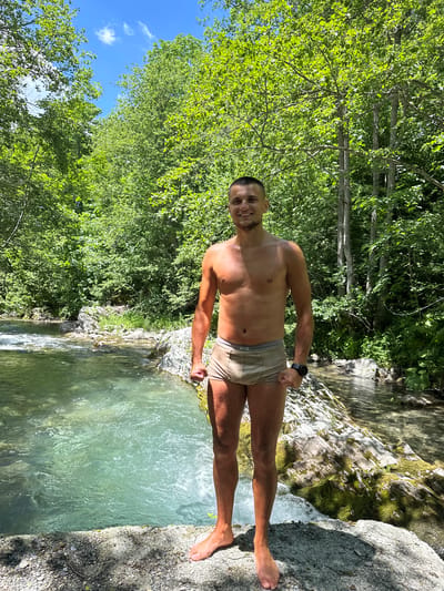 In the city of Bare Kraljske, Montenegro, a young Caucasian male stands shirtless and wearing boxer briefs on a rocky outcrop beside a clear, flowing river. He has short brown hair and a stubble beard, and he is smiling at the camera. His arms are flexed, and he is wearing a black watch on his left wrist. The river is surrounded by lush green trees and foliage, with sunlight filtering through the leaves. The water is a bright turquoise color, and the rocks in the riverbed are visible. The scene suggests a warm, sunny day in a natural, outdoor setting.