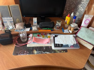 The image depicts a desk surface with various items, including a computer monitor, speakers, pens, a jar, a tube of cream, and other small objects. The items are arranged in a somewhat cluttered manner, suggesting a workspace or study area. The background features a patterned wallpaper and a gold-colored fabric. The overall impression is that of a personal desk with everyday objects. Based on the provided context, this desk is located in Skopin, Russia.