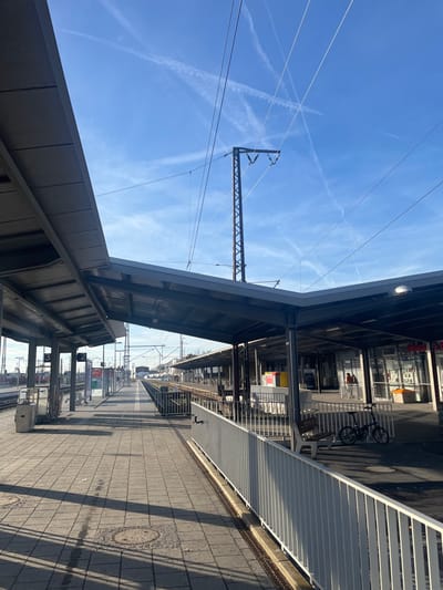 The media file captures a bright, sunny daytime scene at a train station platform in Schweinfurt, Germany. The image is oriented sideways, but should be viewed as if the sky is at the top.

The central focus is a train platform paved with square-tiled stones, stretching into the distance. Long shadows are cast across the platform from overhead structures and buildings, indicating a clear, sunny day. To the left, a modern, grey-paneled canopy covers part of the platform. Above this, the vibrant blue sky is crisscrossed with numerous white contrails from airplanes, forming an "X" shape and other linear patterns. Prominent overhead catenary lines and a sturdy metal support structure for them are visible against the sky.

On the right side of the platform, a white slatted fence or barrier separates it from adjacent buildings. These buildings feature large windows, suggesting shops or station facilities. A black bicycle is parked near a bench in front of these buildings. A person is faintly visible in the background near the bicycle.

On the platform itself, there are various station fixtures, including signs. One clear sign displays the numbers "2" and "3", likely indicating platform or track numbers. Other indistinct signs and information boards are also present. The train tracks are visible in the distance, but no trains or active movement are depicted, suggesting a quiet moment at the station. The overall atmosphere is calm and functional, typical of a train station on a clear day.