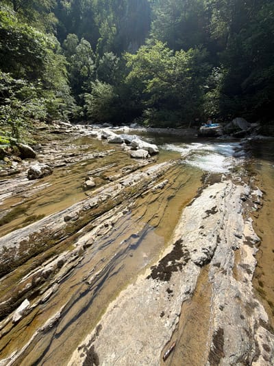 The image depicts a shallow, rocky riverbed with clear, flowing water, surrounded by lush green trees on a sunny day. In the background, two individuals are visible near the riverbank, one sitting on a blue towel and the other standing. The location is identified as Khleborob, Russia. The riverbed is characterized by layered, grey and beige rocks with some moss and dark algae clinging to their surfaces. The water is shallow, allowing the rocks beneath to be clearly seen. Sunlight filters through the dense foliage of the trees, creating dappled light patterns on the water and rocks. The overall scene conveys a natural, tranquil environment.