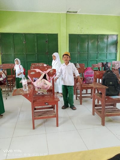 The image captures an indoor scene, identified as a classroom in Jagapura Kulon, Indonesia. The room features light green walls with two sets of dark green, multi-panel wooden doors at the back. The ceiling is white and panelled, showing minor damage near the top left corner. The floor is covered with light-colored square ceramic tiles.
Numerous old, reddish-brown wooden individual desks with attached chairs are scattered throughout the room. A clear plastic "AQVA" water bottle sits on the desk in the left foreground. Three backpacks are visible: a light pink "KUROMI" backpack hangs on a chair in the mid-ground right, another light pink/beige backpack is draped over a central desk, and a dark (possibly black) backpack rests on a desk to the far right. A red backpack is visible on a chair in the far left background.
Three young individuals, approximately 6-9 years old, are prominently featured. In the center-right foreground, a boy stands facing the camera with an open-mouthed expression. He wears a white long-sleeved shirt, dark green trousers, dark sandals, and a yellow cap. To his left, a girl in a white hijab, white long-sleeved shirt, and dark green trousers stands behind a desk, looking to her left. Behind the boy, another girl, also in a white hijab and a long white dress, stands between desks, looking to her right. A partial view of a fourth person, wearing green trousers, is visible on the far left edge of the frame.
