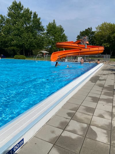 The image captures a bright, sunny day at an outdoor swimming pool complex, likely the public pool in Deutschkreutz, Austria, given the presence of a "1,00m" depth marker. The pool itself is a vibrant, clear blue, with several people visible enjoying the water. In the foreground, the edge of the pool features a white, textured overflow channel and a concrete patio tiled with grey squares, some of which are wet.
Dominating the background and to the right are two large, interconnected orange water slides. A metal railing surrounds the base of the slides and the top platform where a few individuals can be seen. Lush green trees form a dense backdrop to the entire scene, with a clear blue sky overhead. A few picnic tables with a canopy are visible on the grassy area behind the pool. The overall impression is of a lively and recreational public space.