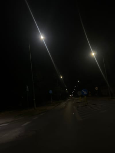 The image depicts a dark, nighttime street scene in Siedlce, Poland, illuminated by streetlights. The camera is positioned low, possibly from the perspective of a moving vehicle or a person walking. A paved road is visible, with white lane markings. On the right side of the road, there are traffic signs, including a blue circular sign with a white arrow pointing right and a smaller circular sign with a red border and a white diagonal bar. Yellow bollards are present near the signs. Trees line the left side of the road, with some foliage visible. The sky is dark and unfeatured. The overall atmosphere is quiet and deserted.