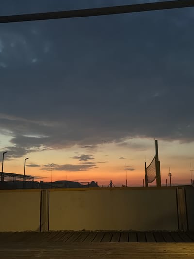 The image depicts a dramatic sunset scene with a mix of dark, cloudy skies on the left and a vibrant orange and yellow gradient on the right, indicating the setting sun. In the foreground, a wooden structure with horizontal planks is visible on the right, suggesting a balcony, deck, or pier. A dark vertical beam runs along the left edge of the image.

In the background, silhouetted structures are visible against the colorful sky. These include what appears to be a large, modern building with a curved roof, possibly a pavilion or shelter, supported by a tall pole. Further back, there are other tall, slender structures that could be lampposts or part of an architectural installation. A faint, dark line in the distance suggests a mountainous or hilly horizon. The sky is filled with scattered clouds, some dark and heavy, others wispy and illuminated by the sunset.

The time of day is clearly sunset, given the intense colors in the sky. The weather appears to be mixed, with both dark clouds and clear, colorful sky. There are no people visible in the image. The overall mood is serene and awe-inspiring, capturing the beauty of nature's light display. There is no visible text in the image.