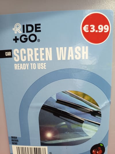 The image shows a close-up of a blue product label for "RIDE + GO SCREEN WASH" which is "READY TO USE". The price tag is a red circle with white text reading "€3.99". The product appears to be for automotive use, with an image of windshield wipers on a car's windshield. The bottom left corner of the label has some numbers: "361034", "08/2025", and "742182", likely a product code and expiry date. There is also a barcode visible. A small graphic of two red tomatoes with green stems is in the lower right corner. The label is likely on a bottle or container. There is no indication of the city of Millstreet, Ireland, or any urban environment.
