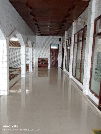 The image shows the interior of a building. The building appears to be a mosque. The building has white tiled walls and a light-brown tiled floor. The floor is wet and shiny, suggesting that it may have recently rained. There is a row of pillars along the left side of the image. The pillars are made of white tile. There is a wooden door at the end of the hallway. The door is closed. The door has a brown wooden frame and a wood-carved design. There are windows along the right side of the image. The windows are made of glass and have a brown wooden frame. The windows have ornate designs etched into the glass. There are no people present. The building is located in Kecamatan Gabuswetan, Indonesia, at the coordinates (latitude: -6.44, longitude: 108.028).