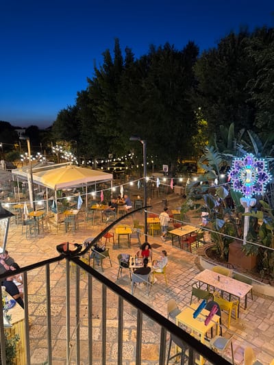 The image shows an outdoor seating area of a restaurant or bar at dusk in Barletta, Italy. The area is paved with light-colored stones and illuminated by string lights and a decorative, illuminated floral motif. Numerous tables and chairs are arranged throughout the space, some occupied by patrons. People are seen dining, conversing, and walking. In the background, trees line the perimeter of the area, and a few cars are visible. The overall atmosphere appears to be casual and lively, with the lighting suggesting it is evening.