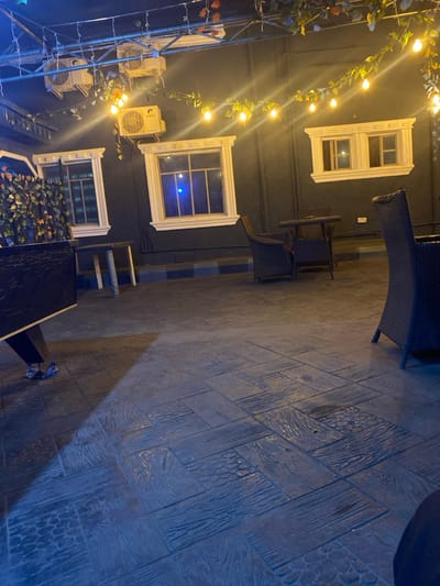 This image depicts an unoccupied, artificially lit outdoor or semi-outdoor seating area at night in the city of Igbessa, Nigeria. The scene features dark walls adorned with two prominent white-framed windows and a smaller pair of white-framed windows above the right one. Two white GREE-branded air conditioning units are mounted on the upper portion of the wall. Overhead, string lights with numerous bulbs are draped, intertwined with artificial green and reddish foliage. A dense screen of artificial foliage is present on the left side of the frame. The floor is paved with dark, textured stamped concrete or tiles designed to resemble wood planks and patterned sections, displaying reflections of the overhead lighting. Furniture includes a partially visible dark wicker chair in the foreground right, a dark rectangular table to the mid-left, and a small dark table flanked by two dark wicker chairs in the mid-center. A horizontal stripe pattern of alternating blue and dark gray runs along the base of the main wall. No individuals are present in the scene.
