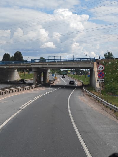 A view from a vehicle traveling on a two-lane asphalt road in Едрово, Russia. The road curves to the right and passes under a concrete overpass with a blue railing. On the right side of the road, there are traffic signs indicating a speed limit of 70, a no parking zone, and a tow-away zone. The environment is rural with trees and grass visible on the embankments. The sky is partly cloudy. Several cars are visible on the road, either approaching or moving away.