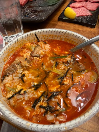 The image captures a dining table scene within a restaurant in the city of Nagano, Japan. In the foreground, a ceramic bowl with an off-white, striped rim contains a red-orange colored soup or stew. This dish includes white rice, pieces of scrambled egg, slices of green onion, dark shredded seaweed, and a visible shiitake mushroom. A silver-colored spoon is partially submerged in the liquid on the right side of the bowl. In the background, to the left, a clear glass tumbler is partially visible. To the right background, a dark, rectangular plate presents several thinly sliced pieces of raw red meat, with a yellow lemon wedge placed alongside. A second dark plate with a green leaf is partially visible further back. The items rest on a visible wooden table surface. No individuals are present in the frame.