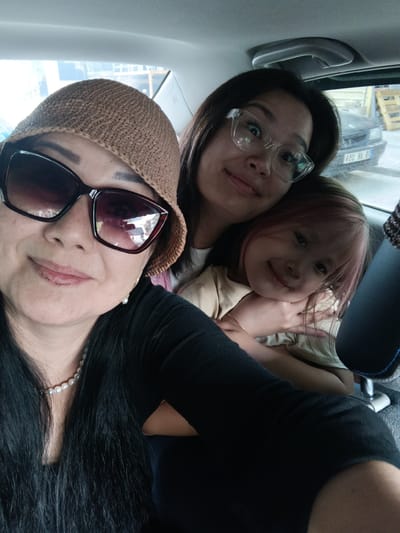In a selfie taken inside a vehicle, three individuals are visible. The person in the foreground, wearing a brown knit hat and large dark sunglasses, appears to be an adult female. She has long black hair and is wearing a pearl necklace. To her right and slightly behind her, a younger female with glasses and dark hair is partially visible. Clinging to the younger female is a small child with pink-tinted hair, also with dark hair. The child is wearing a light-colored t-shirt. The background shows the interior of a car, including a headrest and a window through which a street scene with buildings and a parked vehicle is visible. The license plate of the parked vehicle is partially obscured but appears to read "E20 RA". The scene is likely in Yahşihan, Turkey, given the presence of Turkish text on a sign in the background, which is unreadable. The lighting suggests it is daytime.