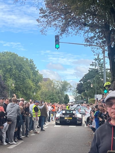A wide shot depicts a black Bugatti supercar proceeding slowly down a paved street in the city of Molsheim, France. A large crowd, estimated at over 100 individuals, lines both sides of the road, attentively observing and photographing the vehicle with smartphones. Several individuals are visible wearing high-visibility yellow or orange vests, positioned among the crowd, suggesting a marshaling or security presence. The Bugatti, an open-top model, displays a yellow British license plate reading "BUG 11." Behind the vehicle, other cars are visible. The environment features mature green trees framing the street, with a clear blue sky and scattered white clouds overhead. A traffic light suspended above the road displays a green signal. In the immediate foreground, a portion of an observer in a grey cap and black hoodie is visible on the right, and other spectators on the left.