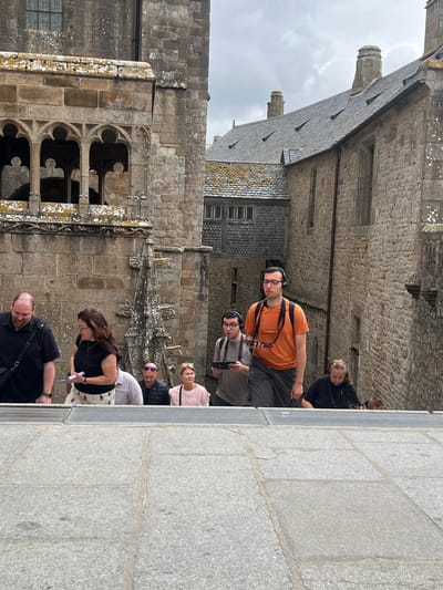 A group of tourists are ascending stone steps in front of a medieval stone building, likely part of Mont Saint-Michel Abbey in Le Mont-Saint-Michel, France. The architecture features stone walls, arched windows, and a slate roof. The sky is overcast. Several individuals are wearing headphones and holding tablets, suggesting they are on a guided tour. In the foreground, large stone slabs form the pathway. The people are diverse in age and gender, dressed in casual outdoor clothing. Some are looking forward, while others are looking down or at their devices.