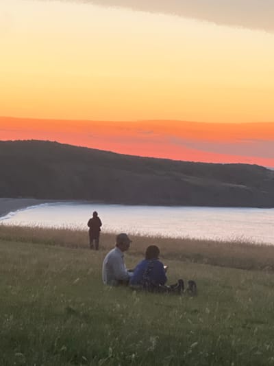 A silhouetted figure stands near the edge of a grassy cliff overlooking a calm body of water. In the foreground, two people are seated on the grass facing away from the camera. One person is wearing a light-colored shirt and a cap, while the other is wearing a blue patterned top. The sky displays a vibrant gradient of orange and yellow, indicating a sunset. The location is the coastal area of Sinemorets, Bulgaria.