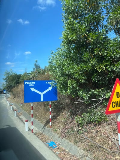 A roadside scene features a blue directional sign with white arrows pointing in three directions, labeled "PLEI KU 40 Km" and "P. KON.". The sign is flanked by red and white striped posts and partially obscured by lush green foliage. Above the sign, a yellow and red triangular sign is visible. Dry grass and a concrete curb line the road, which leads towards the blue sky and scattered clouds.