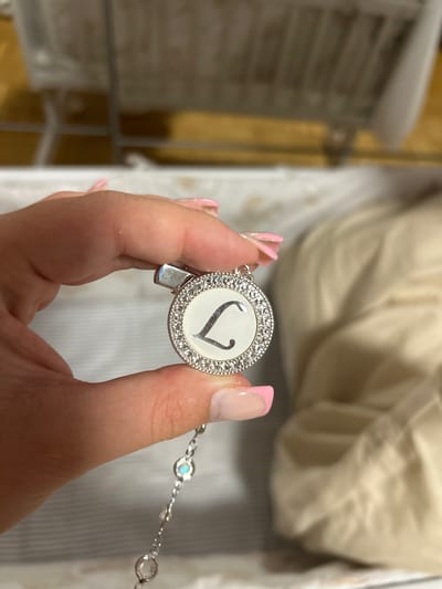 A person's hand with pink-painted nails is holding a round, silver-colored pendant. The pendant has a white face with a black letter "L" in the center. The edge of the pendant is decorated with small, clear stones. A delicate silver chain with additional small clear stones is attached to the bottom of the pendant. The background is blurred, but appears to be an indoor setting with furniture. This scene is not located in Waldbröl, Germany.