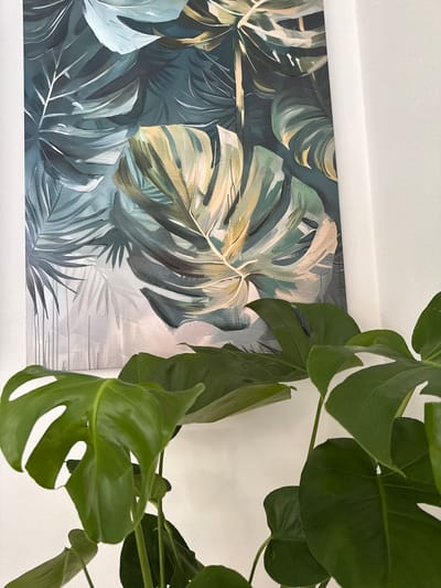 The image shows a close-up view of a Monstera plant in front of a framed picture. The plant features vibrant green leaves with characteristic fenestrations (holes). The framed picture behind the plant depicts a tropical scene with various shades of green and white, showcasing leaves reminiscent of the Monstera plant in front. The background appears to be a white wall. The location is not discernible from the image.
