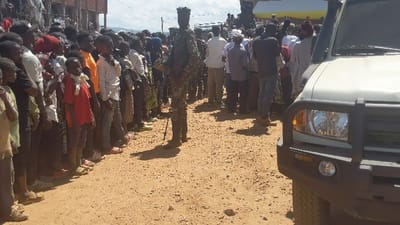 A crowd of people, mostly young, stand together on a dirt road, seemingly observing an event. A uniformed officer stands in the foreground, positioned between the crowd and a white vehicle with a black bull bar. The scene appears to be in a rural setting, possibly a village, with a building visible in the background. The light suggests it is daytime, and shadows indicate the position of the sun.