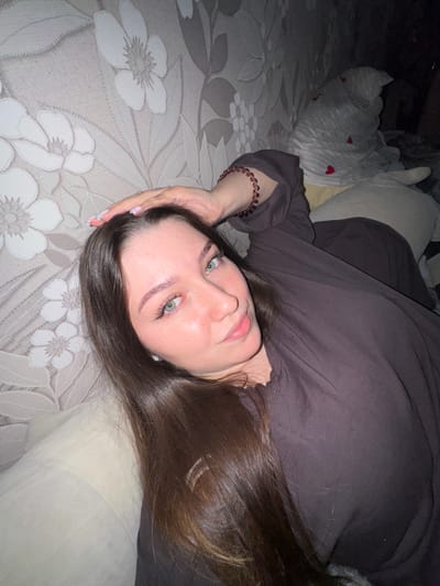 A young woman with long, dark brown hair is lying on her back, propped up against a wall with floral wallpaper. Her right hand is resting on her head, and she is wearing a dark, long-sleeved shirt and a reddish beaded bracelet on her left wrist. She has light-colored eyes and is looking towards the camera with a slight smile. In the background, partially obscured, is a white stuffed animal with small red hearts. The overall lighting suggests an indoor setting, possibly a bedroom, in Shirkovo, Russia.