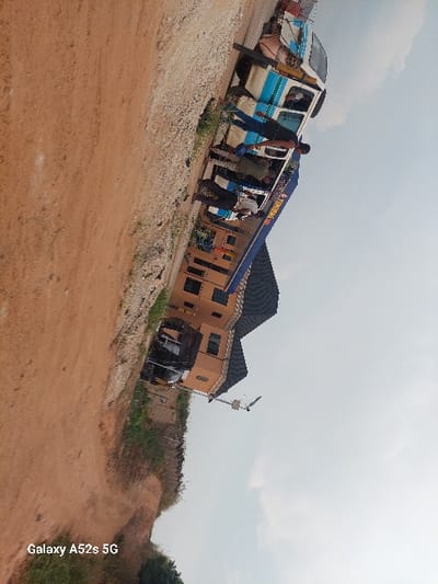 A slightly tilted view captures a partially damaged blue and white bus on a dusty road, with passengers disembarking. Adjacent to the bus is a yellow building with a dark roof, featuring visible windows and a black vehicle parked nearby. The background includes sparse greenery, a utility pole with a satellite dish, and a cloudy sky. The dusty road leads to a higher elevation on the right side of the frame.