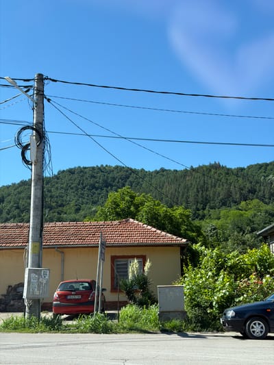 The image depicts a residential street scene in the village of Beli Osam, Bulgaria, under a clear blue sky. A concrete utility pole, heavily laden with black electrical wires, stands prominently on the left side of the frame. To the right of the pole is a yellow house with a red-tiled roof. A red hatchback car is parked in front of the house, displaying the license plate "OB 6357 BB". Adjacent to the car and house is a dense area of green vegetation and trees. In the background, a lush, tree-covered hillside dominates the landscape. On the right side of the image, the front of a dark-colored car is visible, parked on the paved road. The road itself shows asphalt markings.