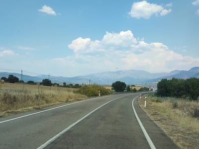 This is a horizontal, eye-level, wide-angle photograph captured on a sunny day, likely in the late morning or early afternoon. The image depicts a winding paved road with white lane markings, curving to the right and leading into the distance. The road is bordered by dry, golden grasses and vegetation on both sides. In the background, rolling hills and distant mountains are visible under a bright blue sky dotted with white, fluffy clouds.
The scene suggests a rural or semi-rural location in Zarza de Granadilla, Spain, given the landscape of agricultural fields, scattered trees, and power lines. Specifically, to the left of the road, several large, round hay bales are visible in a dry field, a common sight in agricultural areas. On the right side of the road, there is a white sign with black lettering that appears to be directional, pointing towards "DOSFAS". Another sign, red and white, shows the number "70", possibly indicating a speed limit.
There are no people visible in the image. The overall mood is serene and peaceful, evoking a sense of travel and open road. The bright sunlight and clear sky indicate fair weather, suitable for driving or outdoor activities. The composition emphasizes the expansive landscape and the journey ahead.