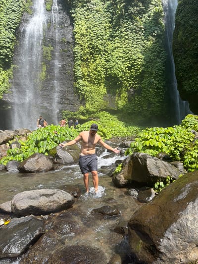 The image depicts a man standing in a shallow river with a waterfall in the background. The man is shirtless, wearing dark shorts and a baseball cap, and holding a selfie stick. He is looking to his left with his arms outstretched. Several other people are visible in the background near the waterfall. The waterfall cascades down a rocky cliff face covered in lush green vegetation. The riverbed is rocky, with clear water flowing over submerged stones. The scene appears to be a natural outdoor environment, likely a tourist attraction given the presence of people and the selfie stick. This location is identified as Sekumpul Waterfall in Sawan, Buleleng Regency, Bali, Indonesia.