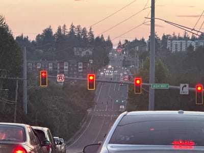 The image depicts a street scene at dusk in North Lynnwood, United States. A traffic light with a red arrow pointing left is visible in the foreground, accompanied by a "no U-turn" sign. Further down the road, a series of traffic lights are red, indicating a stop. Several cars are lined up, waiting at the intersection. The background shows a residential area with apartment buildings and trees lining a hilly landscape. The sky is a pale orange and pink, characteristic of sunset. The overall atmosphere is calm and orderly, with traffic flowing smoothly despite the red lights.