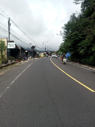 The scene depicts a street view in Saing Ta Mawt Lay, Myanmar, under an overcast sky. A dark asphalt road, marked with a solid yellow line on its right side and a broken white line on its left edge, extends into the distance, curving slightly to the right.
On the left side of the road, a concrete shoulder abuts a series of structures. Closest is a building featuring a sign that reads "BENGKEL MOBIL" with an "MG" logo, indicating a car repair shop. Adjacent to this, a utility pole is heavily laden with numerous black electrical and communication wires. Beyond this, a row of single-story commercial buildings or residences, some with corrugated metal roofs, line the street. Several individuals are visible on the sidewalk area, with one person in a light blue shirt standing near the "BENGKEL MOBIL" sign. A yellow utility truck or small bus is parked or moving slowly further down the road on this side, followed by a white SUV.
The right side of the road is bordered by a dense, verdant expanse of trees and foliage, indicative of a natural, possibly sloping, area. A partially obscured blue tarp is visible within the vegetation. A yellow diamond-shaped road sign with a black arrow is present further down this side.
Vehicles are present on the road. In the mid-ground, a person wearing a dark helmet and clothing is riding a light-colored scooter, appearing to move away from the viewer. Other motorcycles and scooters are visible both ahead of and behind this primary scooter, generally traveling in the same direction.