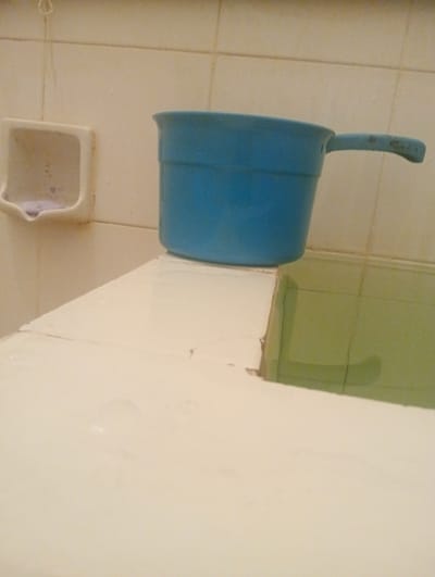 This image shows a bathroom scene. The primary objects visible are a blue plastic dipper with a handle and a white soap dish mounted on a tiled wall. The dipper is placed on a white ledge that appears to be the edge of a bathtub or sink. The background consists of light-colored, square tiles on the wall. Below the white ledge, there is a green tiled area, likely part of the bathtub or shower. A small piece of soap is visible in the soap dish. The lighting suggests it is daytime, and the overall impression is of a functional, somewhat dated bathroom. No people are visible, and no text is present in the image. The location context is Surabaya, East Java.