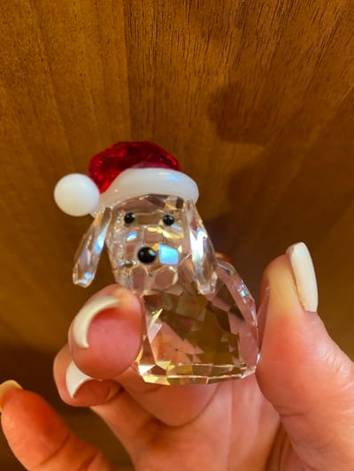 A hand with white-painted nails holds a small, faceted glass figurine of a dog wearing a Santa hat. The figurine has black eyes and a red hat with a white trim and pom-pom. The background is a warm-toned wooden surface, likely a table. The lighting appears to be indoor, creating reflections and refractions within the glass object. The hand is positioned to cradle the figurine, suggesting a moment of appreciation or display. No text is visible in the image. The setting is likely a home or indoor space given the wooden surface and close-up nature of the shot.