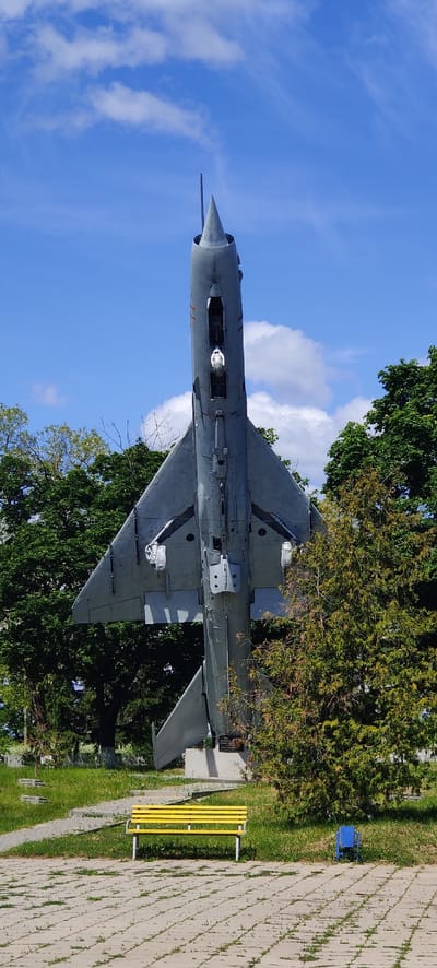A MiG-21 fighter jet is displayed on a concrete plinth in Kolomak, Ukraine. The aircraft is painted in gray camouflage. A yellow bench is located nearby. Lush trees surround the aircraft, partially obscuring it from view. A blue sky with white clouds forms the backdrop. A paved walkway is visible to the right.